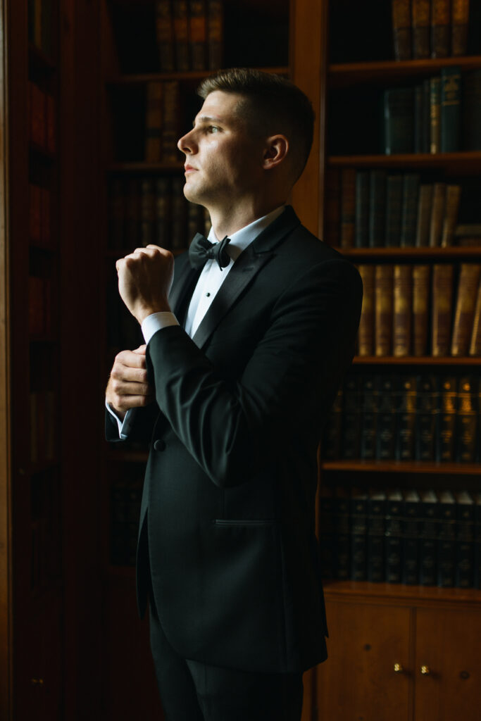 Groom fixing his cuff link while in the library of boxwood estate on his wedding day morning