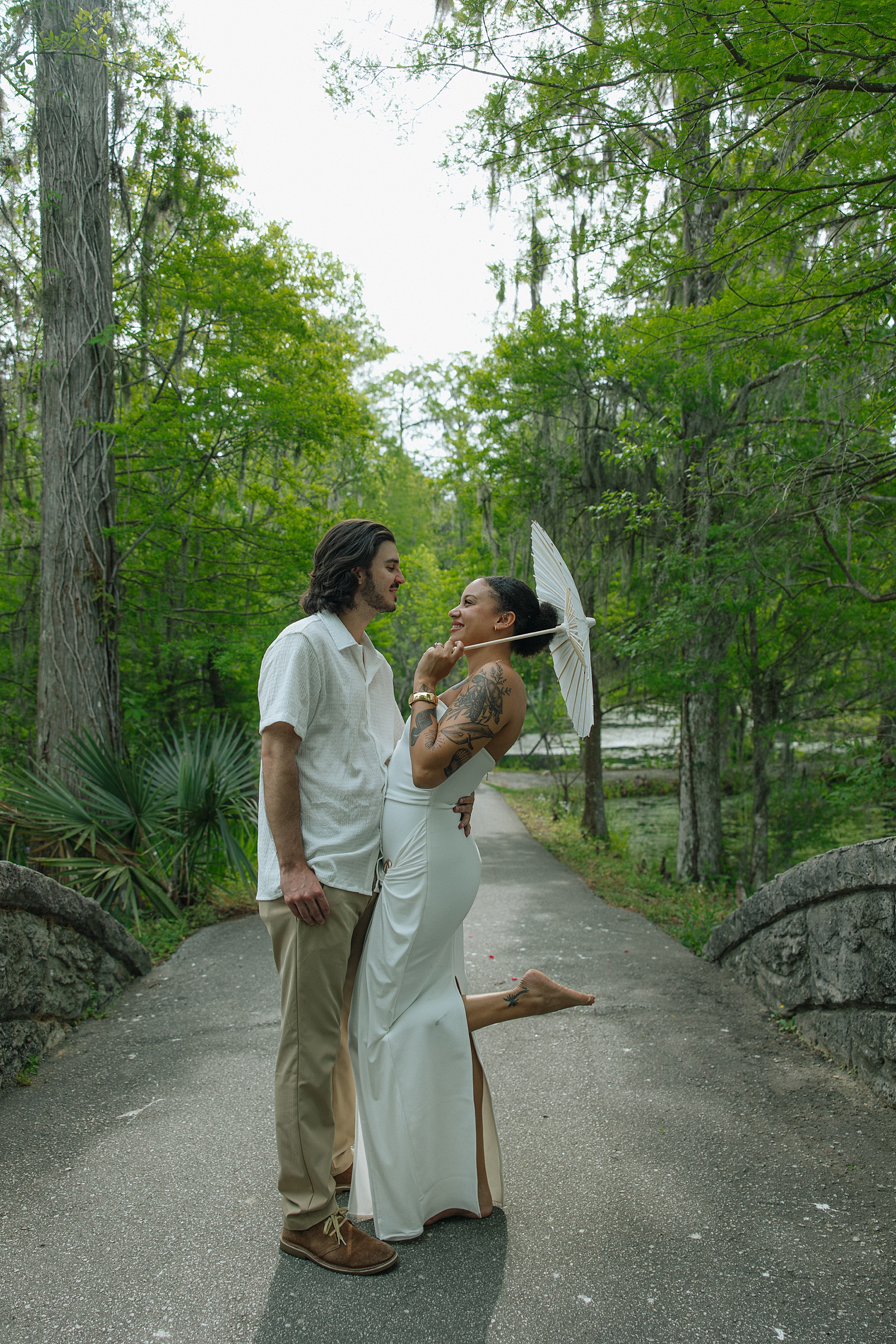 A happy engaged couple stand in a park sidewalk in white holding each other and a parasol