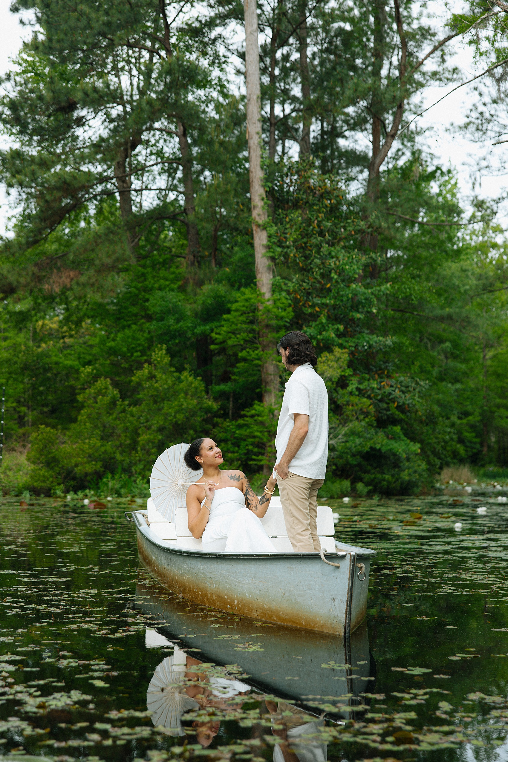 A man stands in a small metal boat holding the hand of his new fiancee in a white dress holding a parasol on her shoulder during their cypress gardens engagement photos