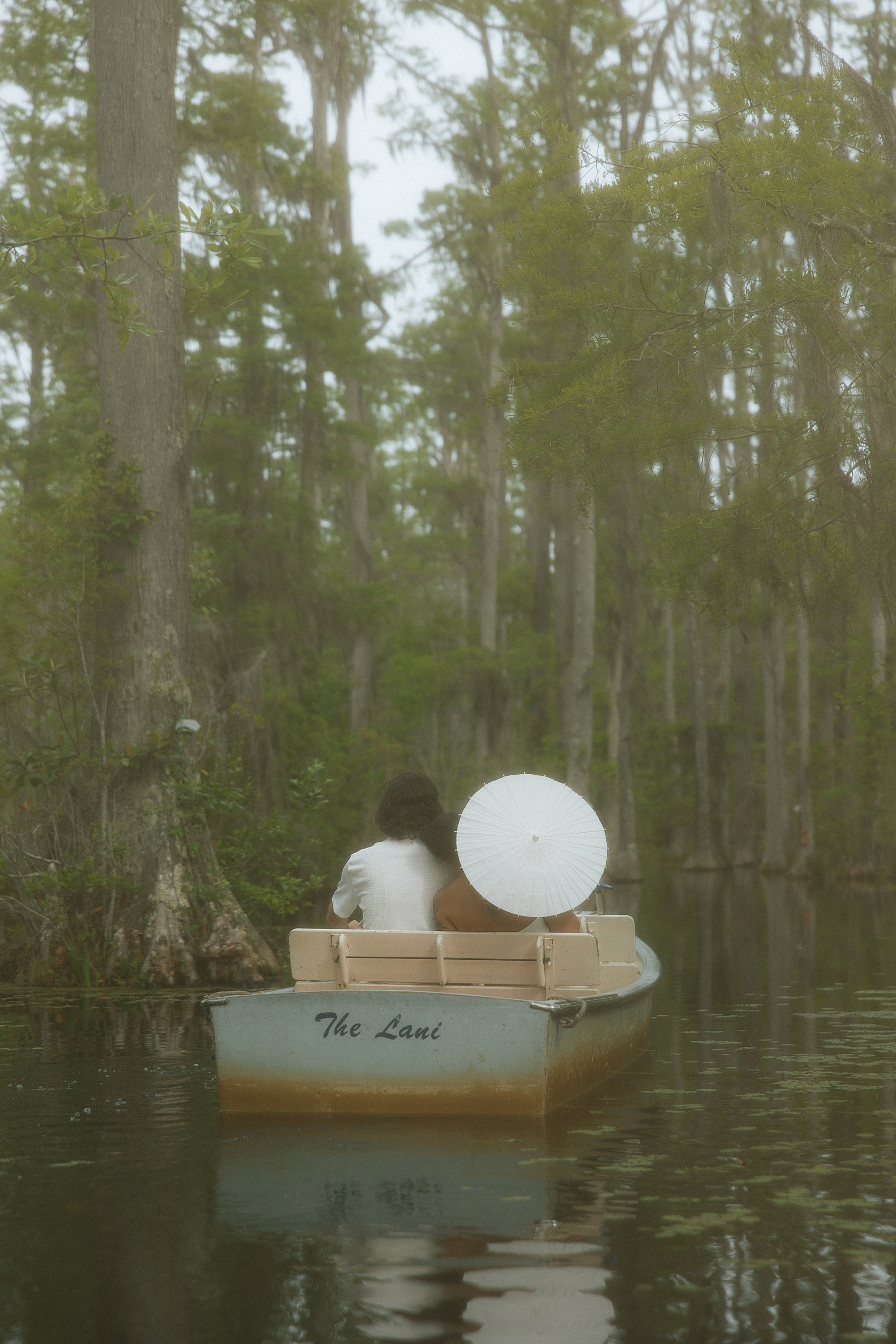 A look at the back of a couple floating in a cypress swamp in white outfits with a matching parasol in a metal boat